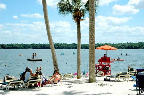 Students on the sandy beach at Lake Wauberg where they are enjoying the water with a life guard in a red life guard chair overlooking to ensure safety
